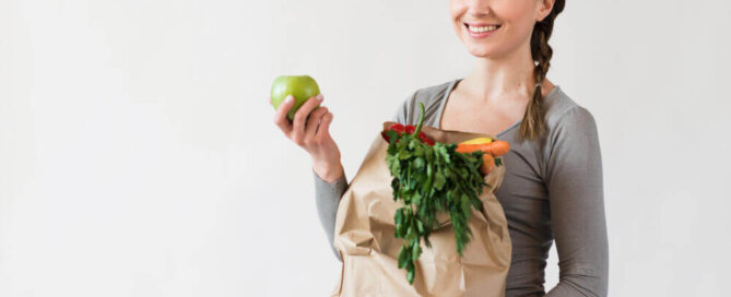 Moça sorridente com uma maçã na mão e com uma sacola de papel reciclado com verduras e frutas, na outra mão