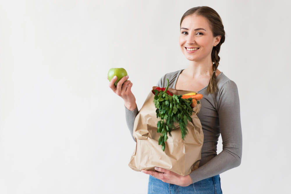 Moça sorridente com uma maçã na mão e com uma sacola de papel reciclado com verduras e frutas, na outra mão